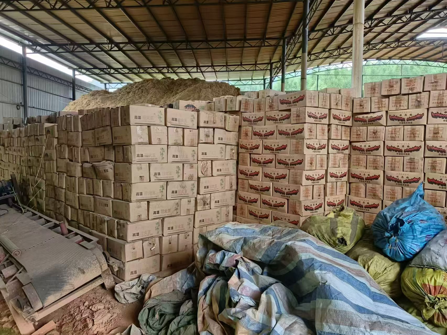 Covered warehouse with stacked cartons of bamboo charcoal briquettes and a pile of bamboo material behind them